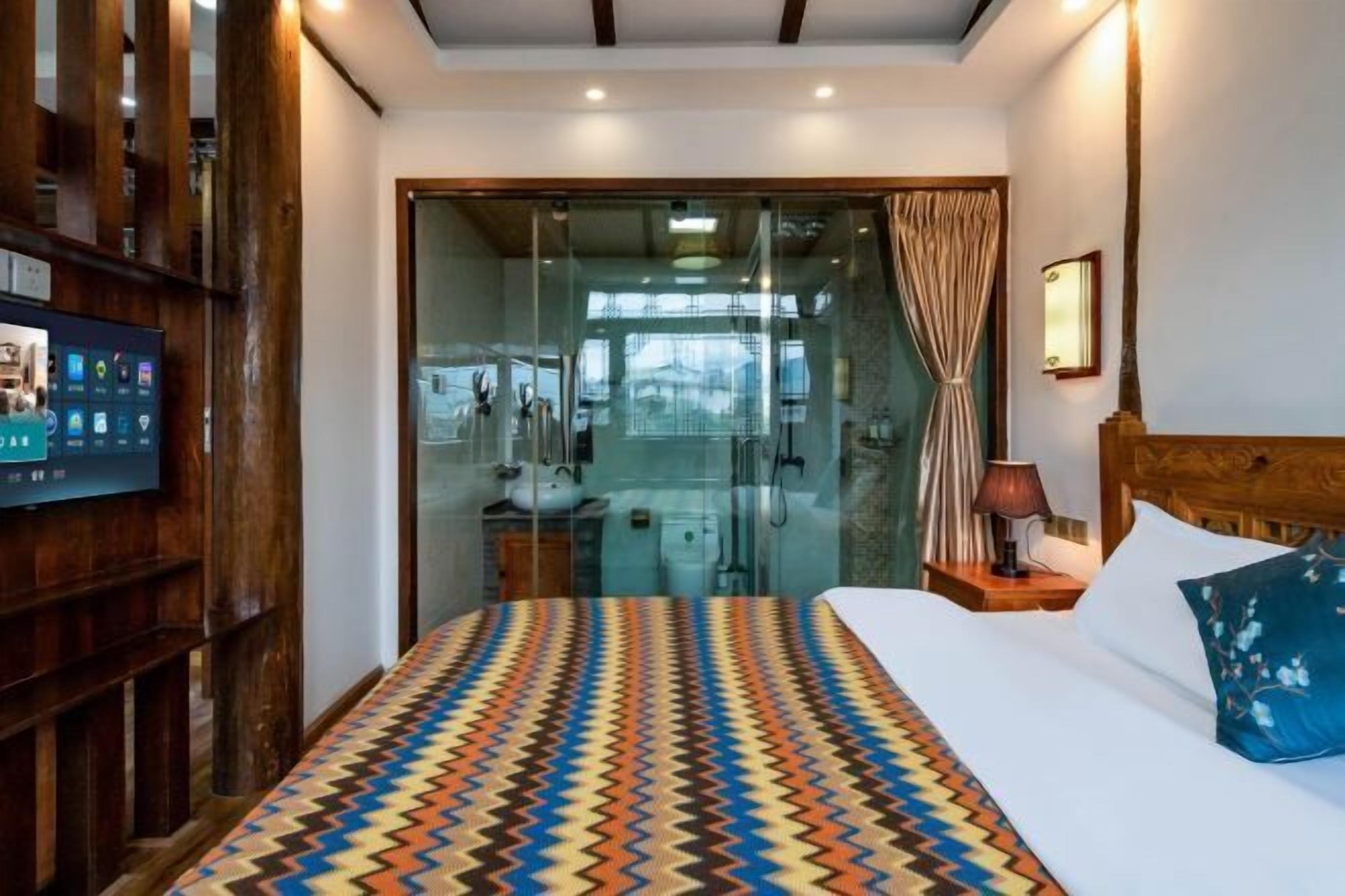 Quarto de hotel com cama, televisão, cabeceira de madeira, mesa de cabeceira e banheiro visível através de uma porta de vidro.