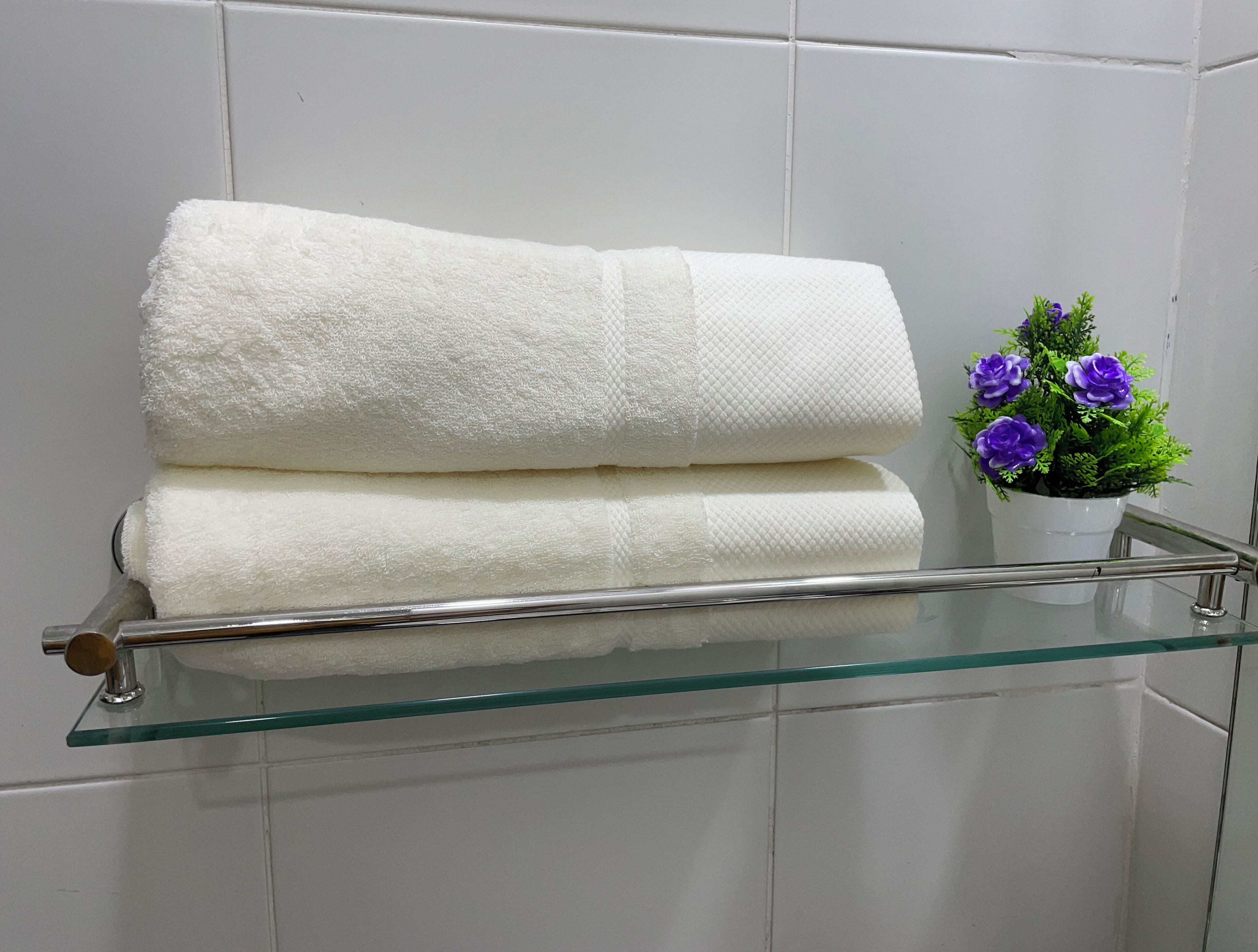 Une étagère en verre sur laquelle sont pliées des serviettes blanches, et une plante en pot aux fleurs violettes.