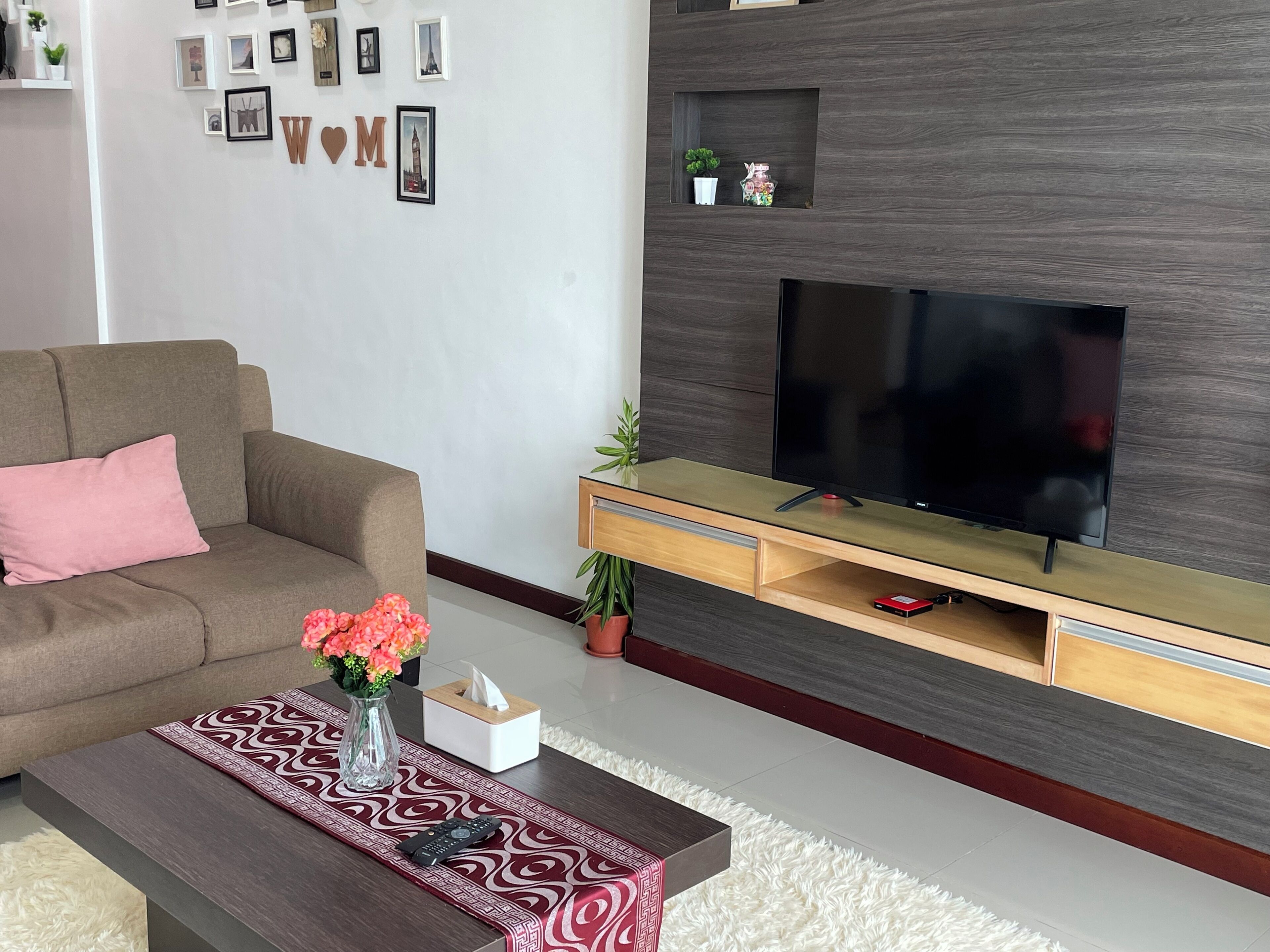 Una sala de estar moderna con un sofá marrón, una mesa de centro con un jarrón de flores, un televisor sobre un mueble de madera, y una pared decorada con marcos de fotos y letras.