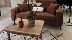 Een moderne woonkamer met een bruine bank, een houten salontafel en een kamerplant. Achtergrond toont een keuken met een eethoek.