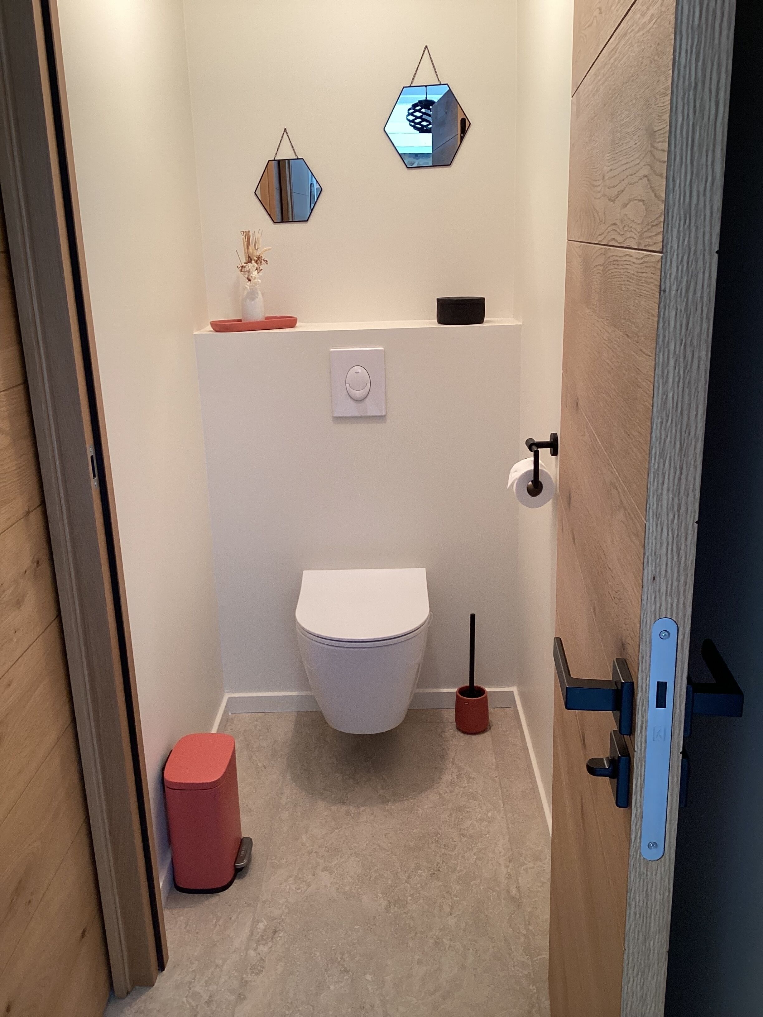 A compact bathroom with a white toilet, a red trash bin, a toilet brush, and two hexagonal mirrors on the wall.
