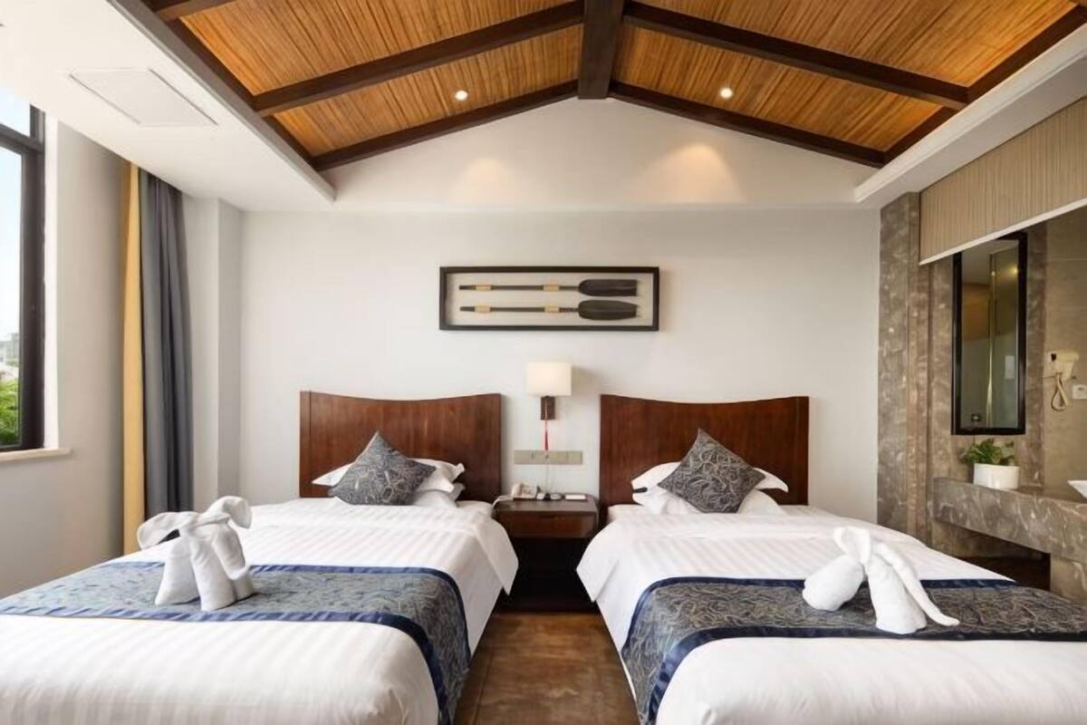 Habitación de hotel con dos camas, techo de madera, mesita de noche con lámpara y vistas al exterior.