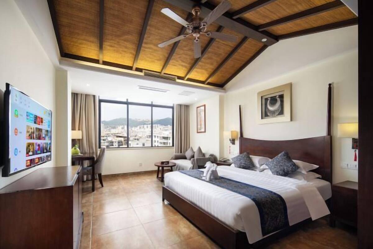 Habitación de hotel con una cama grande, un escritorio, un televisor y vistas a un paisaje urbano a través de la ventana.