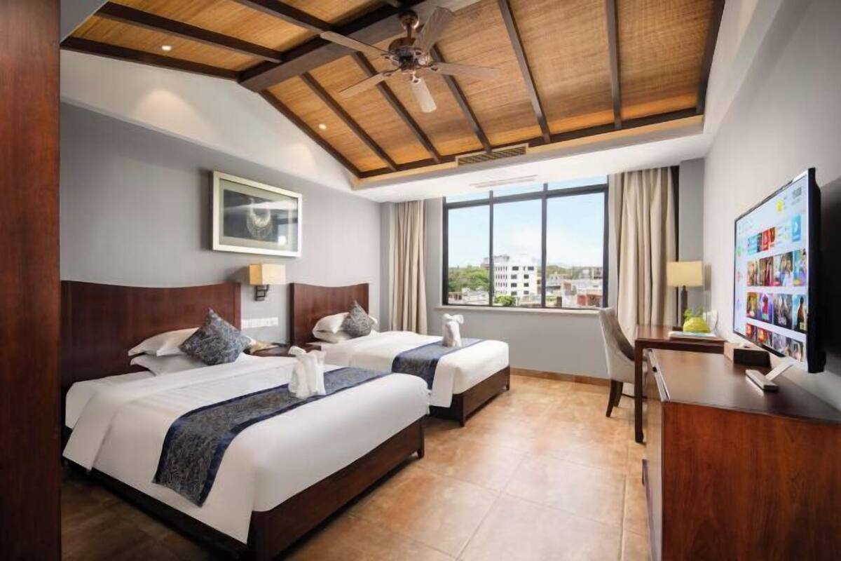 Habitación de hotel con dos camas, un escritorio, un televisor y un ventanal con vista a la ciudad.