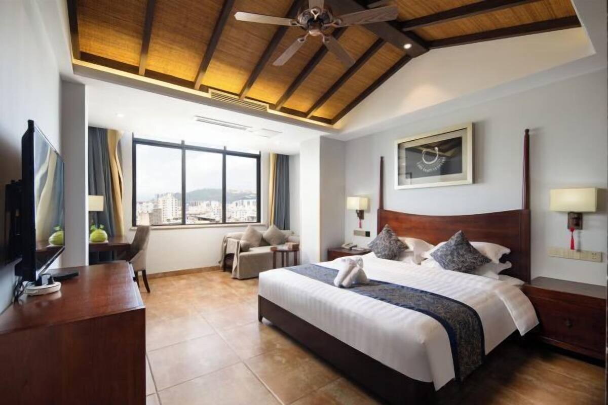 Habitación de hotel con cama, escritorio, silla, televisión, ventilador de techo y un gran ventanal.