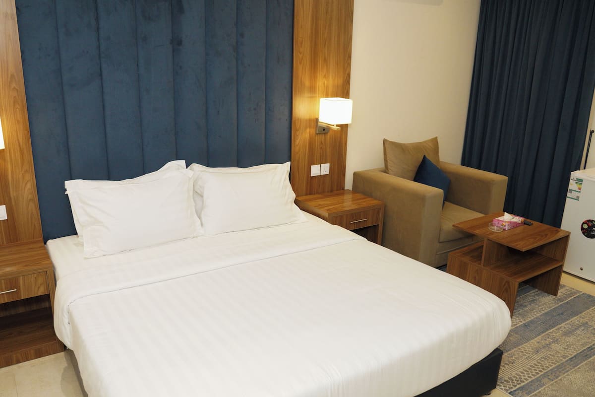 A hotel room with a large bed, a wooden nightstand, a desk, a chair, and a small refrigerator.