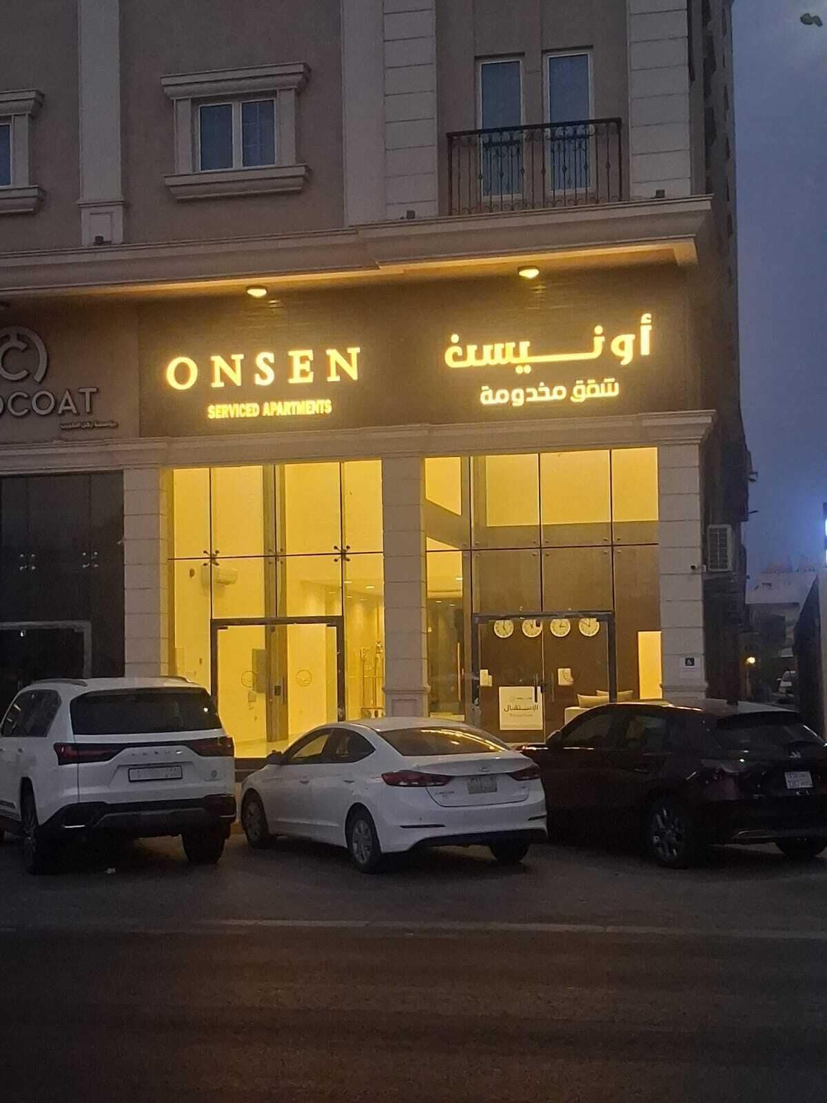 A building with a sign for 'ONSEN Serviced Apartments' and Arabic text. There are three cars parked in front.