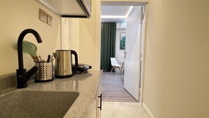 A narrow hallway with a kitchen area on the left, including a sink, a kettle, and a utensil holder. A chair and a table are visible in the background. - AQ 21 New Studio with kitchen and self check-in (Riyadh)