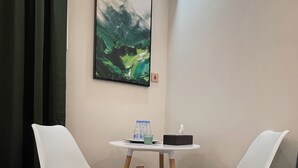 A room with two white chairs, a small table with a tissue box and water bottles, a gray sofa, an air conditioning unit, and a painting on the wall. - AQ 21 New Studio with kitchen and self check-in (Riyadh)