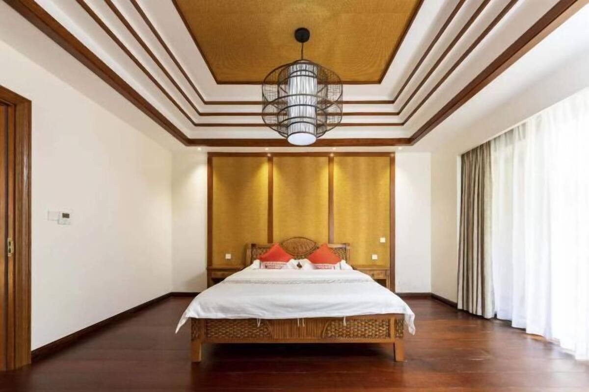 A room with a bed, a wooden floor, a ceiling with a patterned design, and a hanging light fixture.