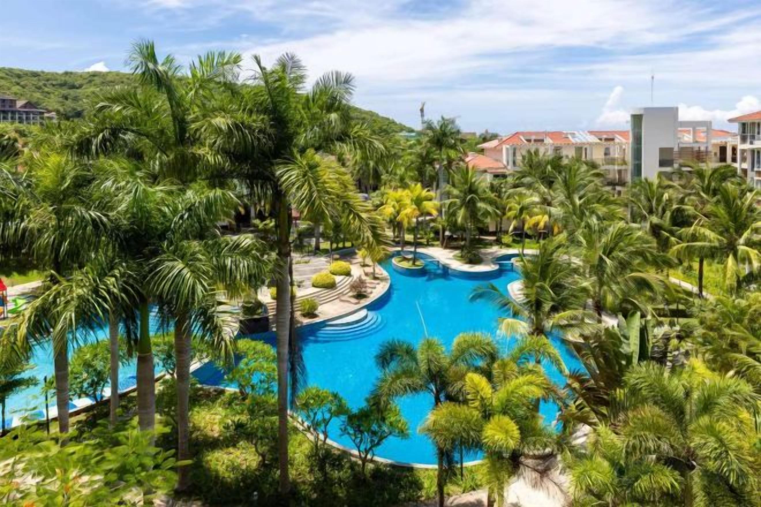 A resort with a large central pool surrounded by palm trees and a mountainous backdrop.