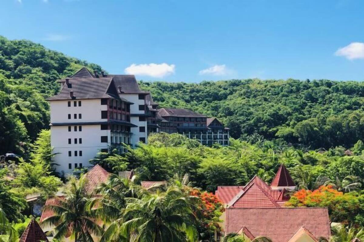 A large building with multiple floors and a steep roof, surrounded by lush greenery and palm trees.