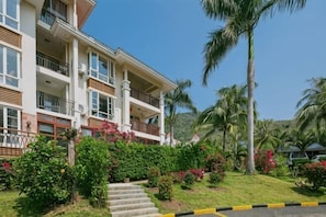 Um hotel com vários andares, varandas, uma escadaria que leva à entrada e paisagismo tropical.