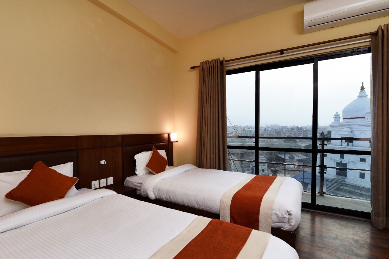 A hotel room with two beds, a large window with a view of a cityscape, and an air conditioning unit on the wall.