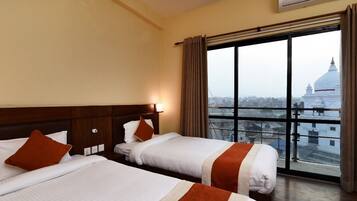 A hotel room with two beds, a large window with a view of a cityscape, and an air conditioning unit on the wall.