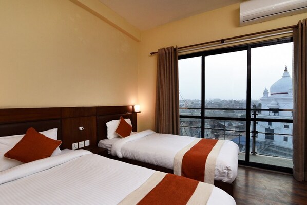 A hotel room with two beds, a large window with a view of a cityscape, and an air conditioning unit on the wall.