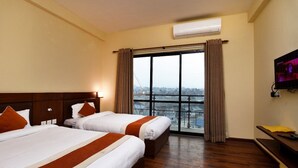A hotel room with two beds, a large window with curtains, an air conditioning unit, and a television on the wall. - Hotel Harmony (Kathmandu)