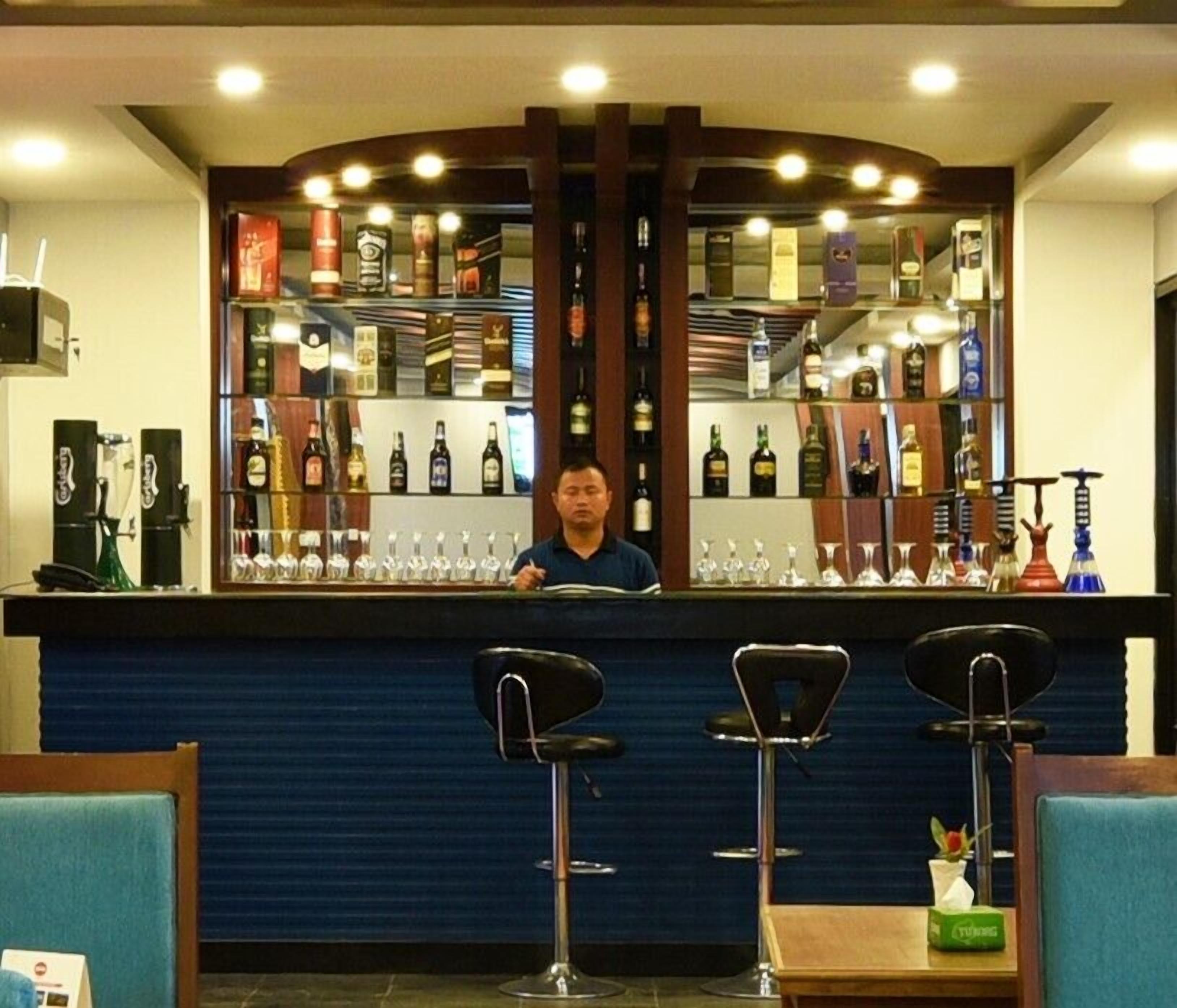 Zona de bar con un camarero, una barra con diferentes botellas y tres taburetes.