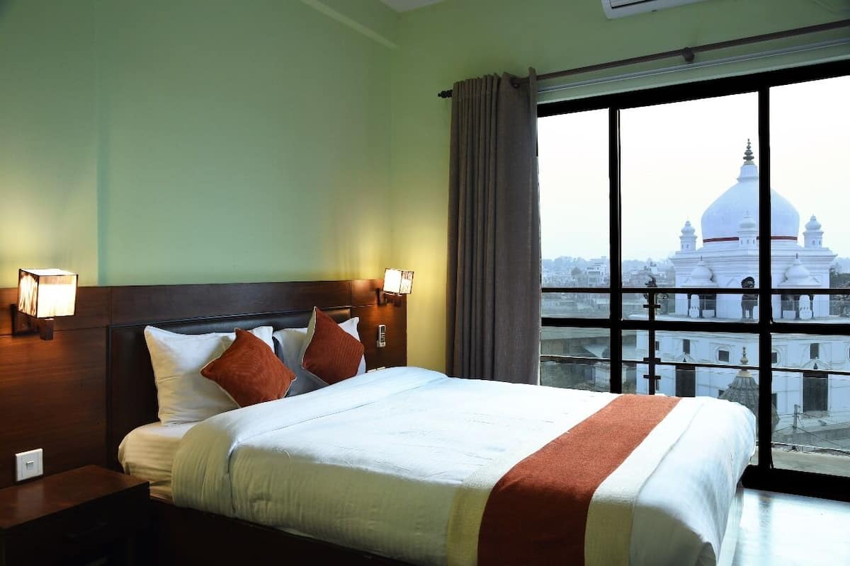 A hotel room with a large bed, two bedside lamps, a nightstand, and a view of a white building with domes through a large window.
