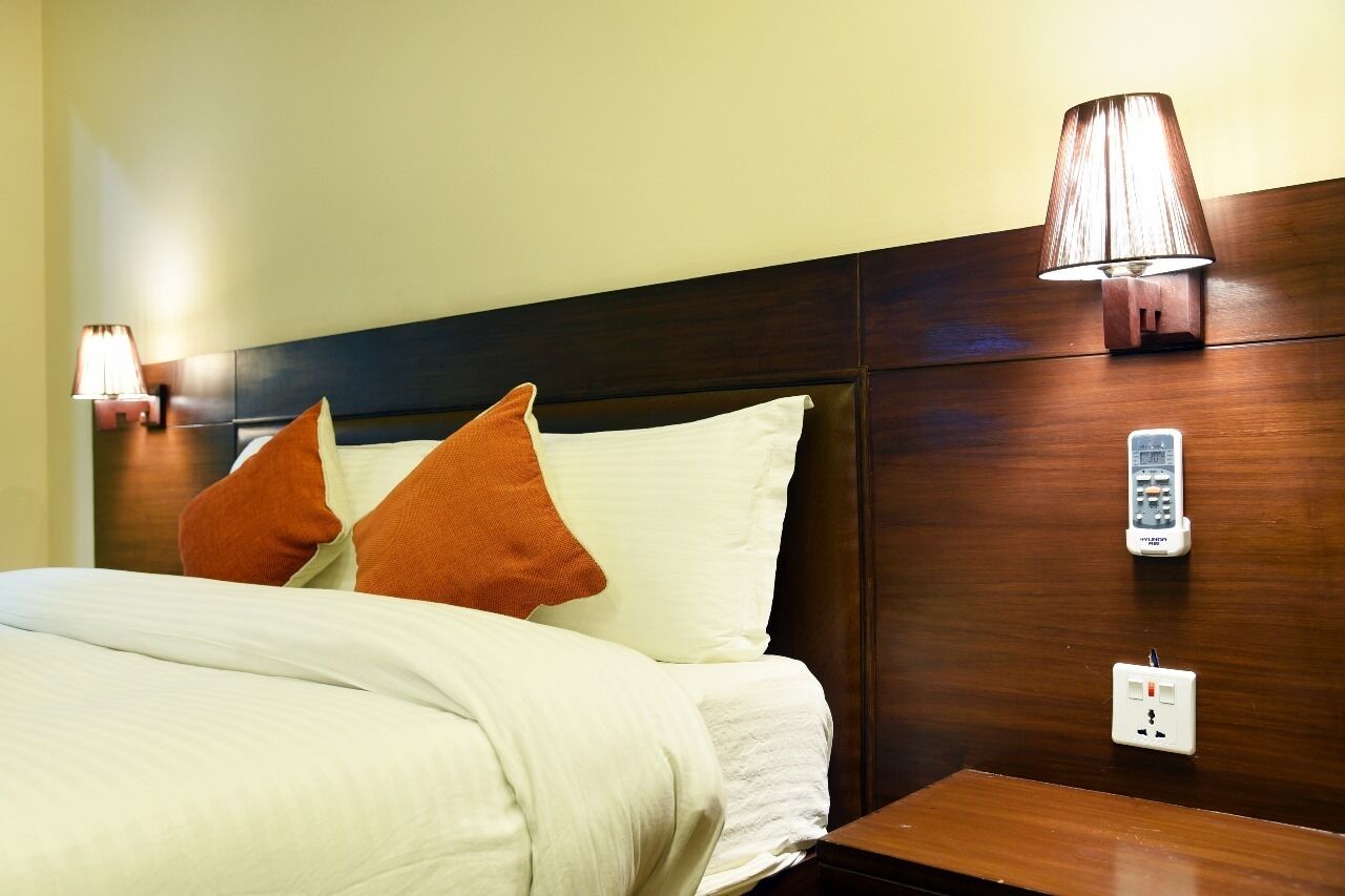 A hotel room with a bed, two pillows, a bedside lamp, a phone, and a power outlet.