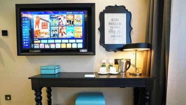 A hotel desk with a TV displaying a movie selection, a framed quote, a lamp, and a small table with a blue box and a cup. - Xana Hotelle (Beijing Changping Metro Station, China University of Petroleum‑Beijing) (Beijing)