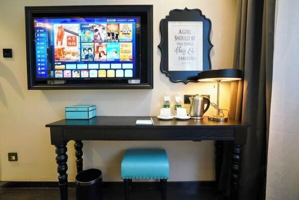 Un bureau d’hôtel avec un téléviseur diffusant une sélection de films, un cadre contenant une citation, une lampe et une petite table sur laquelle se trouvent une boîte bleue et une tasse.