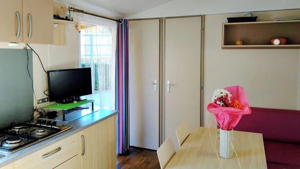 A compact kitchen with a stove, a television on a stand, a dining table with chairs, and a sofa. - Seasonal Mobile Home (Les Arcs)