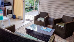A patio with wicker furniture, a glass table, and a view of a house through a sliding door. - Seasonal Mobile Home (Les Arcs)