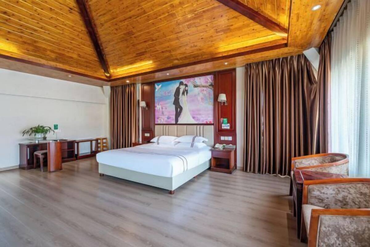Quarto de hotel com uma cama grande, teto de madeira, uma escrivaninha com cadeira, uma tela de TV e uma cortina.