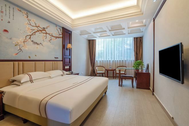A hotel room with a large bed, a desk with two chairs, a TV, and a decorative wall with a painting.