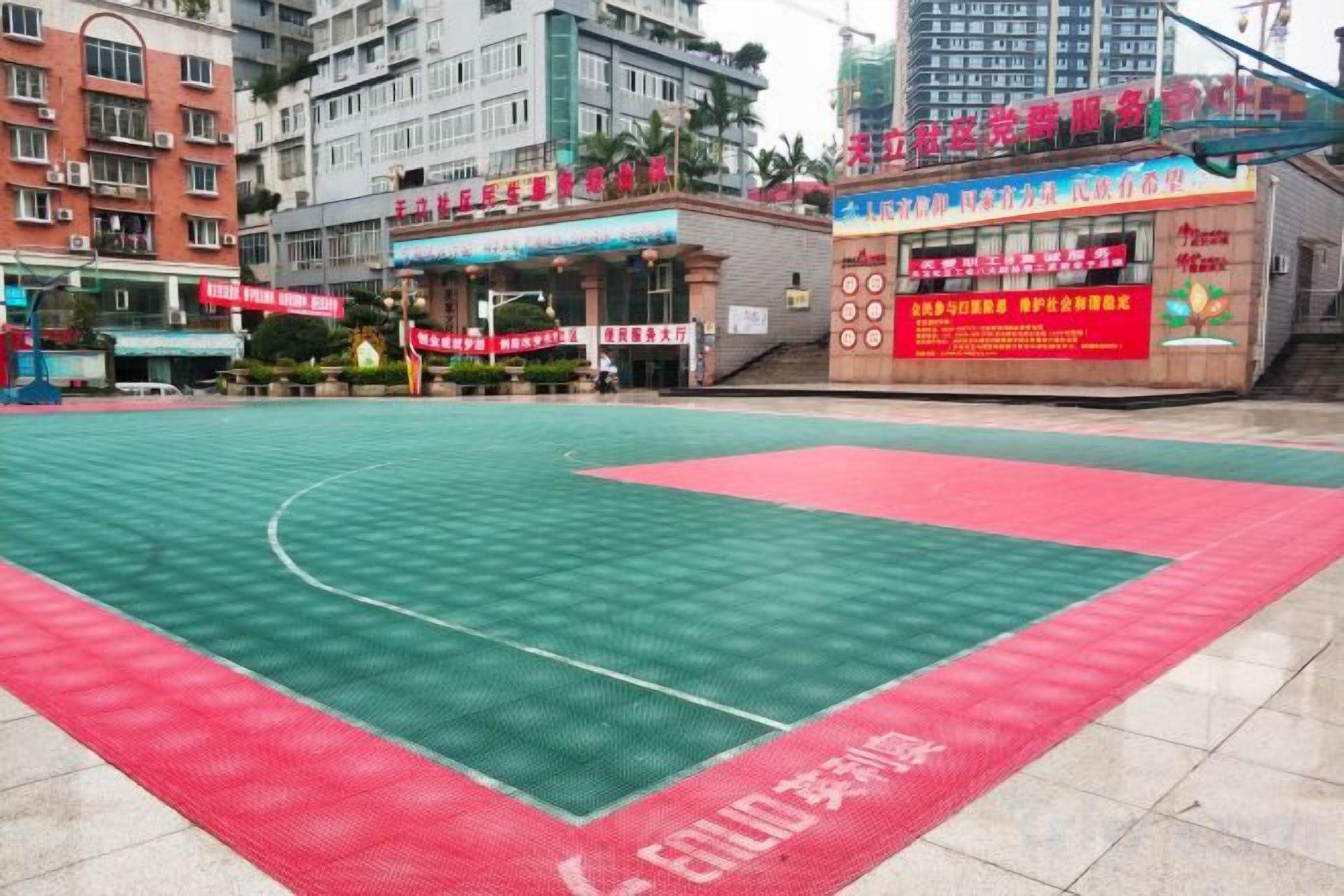 An outdoor basketball court with a red and green surface, surrounded by buildings and a food stall.