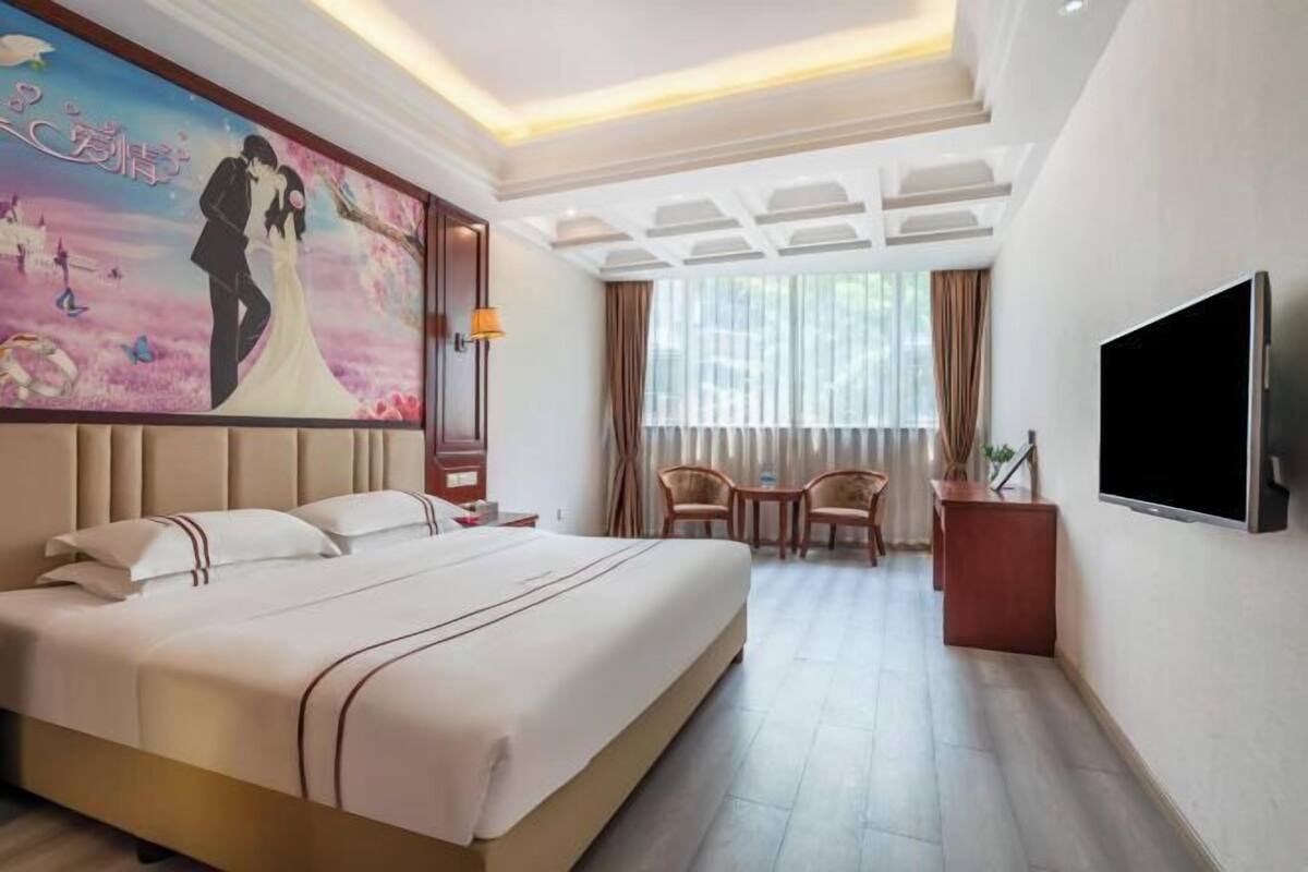 Quarto de hotel com uma cama grande, televisão, escrivaninha com cadeiras e janela com cortinas.