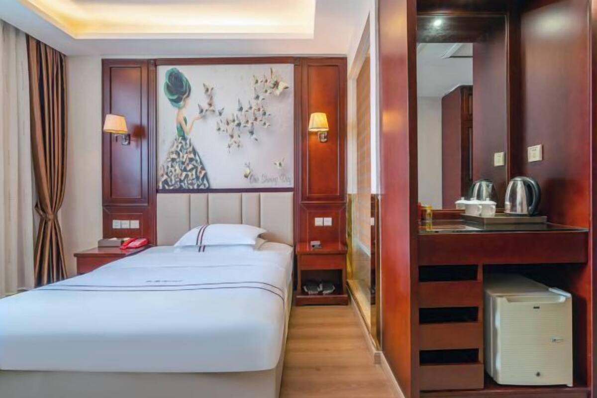 Quarto de hotel com uma cama grande, uma escrivaninha com chaleira, um pequeno frigorífico e um quadro na parede.