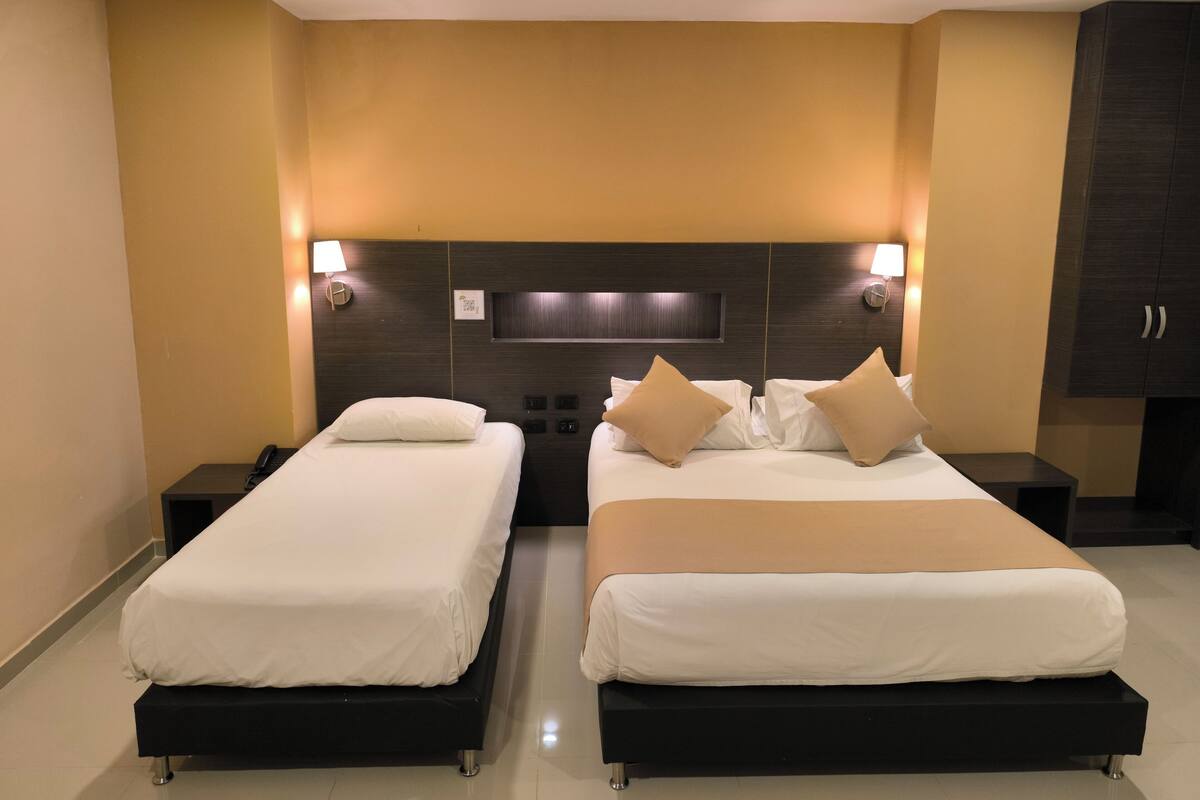 A hotel room with two single beds, a large bed, a dark wood headboard, wall-mounted lights, and a dark wardrobe.