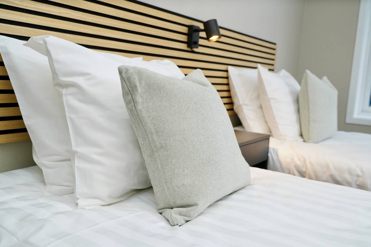 Un lit bien fait, avec une literie blanche et une variété d’oreillers, dont un oreiller gris à texture particulière.