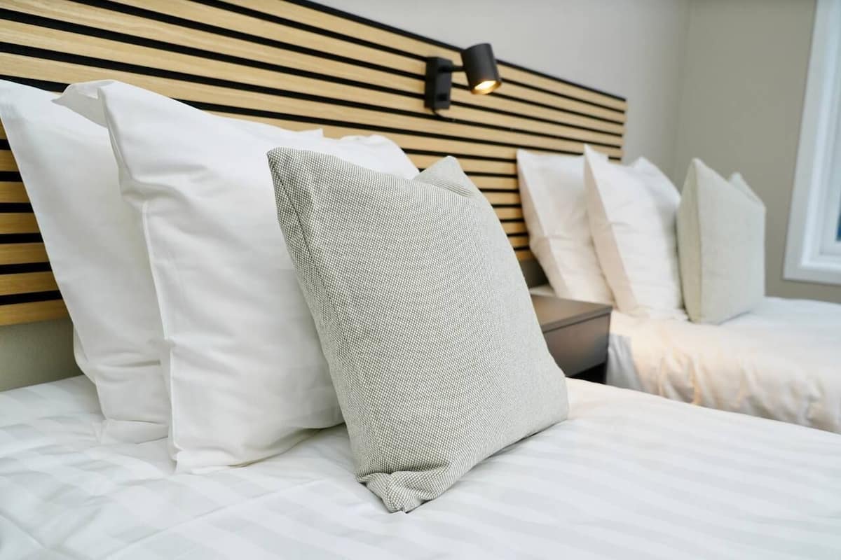 Un lit bien fait, avec une literie blanche et une variété d’oreillers, dont un oreiller gris à texture particulière.