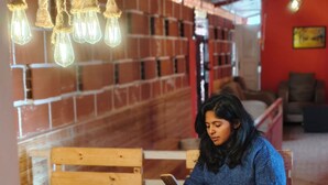 Une personne est assise à une table en bois dans une pièce aux murs de briques, où sont suspendues des ampoules.