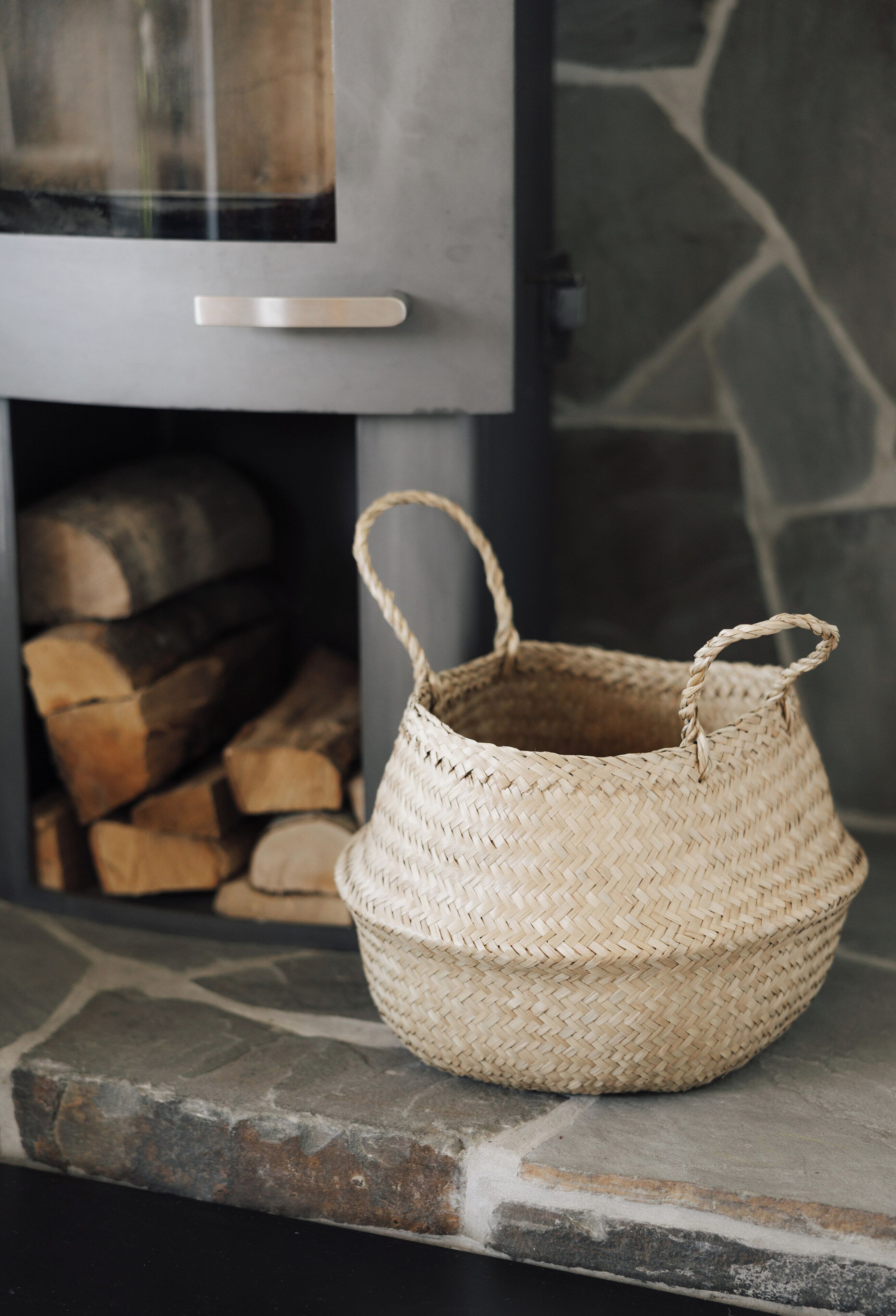 Una cesta tejida con asa sobre un repisa de piedra cerca de una chimenea con leñera.