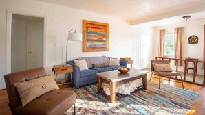 A living room with a blue sofa, a brown leather chair, a coffee table, and a colorful rug. There is a painting on the wall and a dartboard on the right. - Urban Haven -Glen Iris/2 BR/Pets/WiFi/Full Kitchen (Birmingham)
