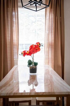A dining table with a vase of red flowers, a modern chandelier, and sheer curtains.