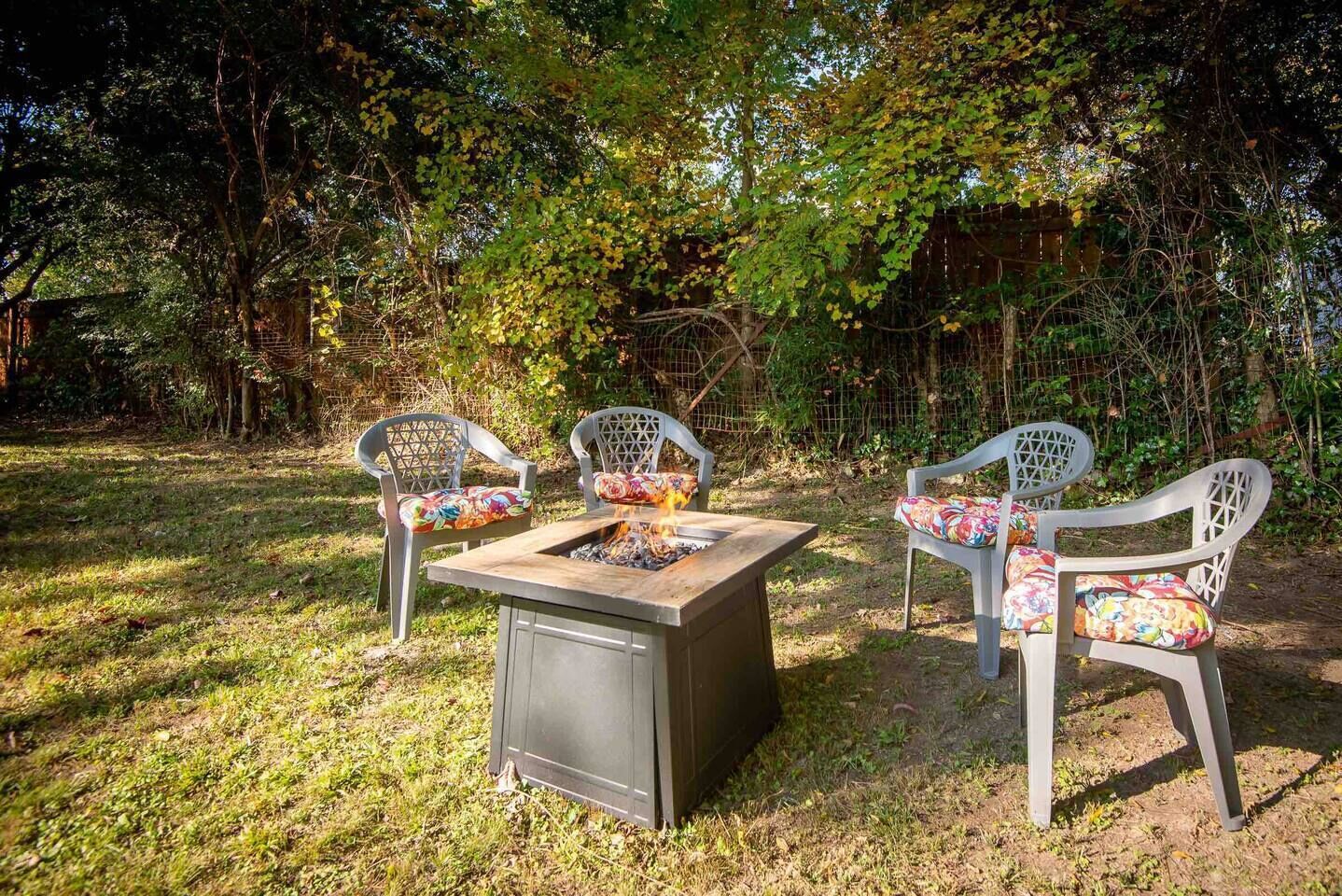 An outdoor seating area with a metal table and chairs, surrounded by trees and a fence.