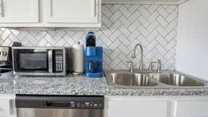 Una cocina con gabinetes blancos, microondas, cafetera, lavavajillas y un lavamanos doble con accesorios de acero inoxidable.