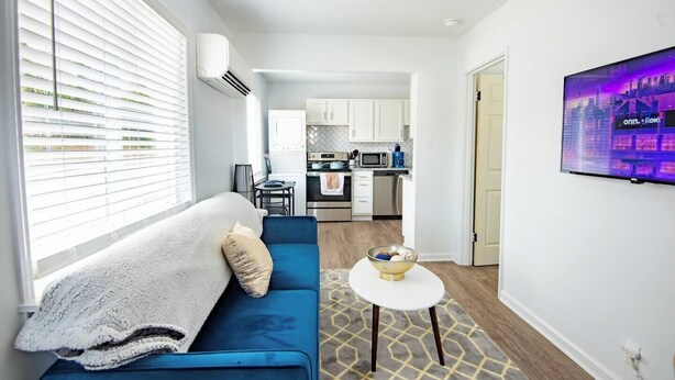 Una sala de estar moderna con un sofá azul, una mesita blanca y una cocina al fondo.
