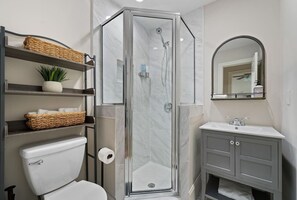 Un baño moderno con ducha acristalada, inodoro, lavamanos y estantes con cestas y una planta.