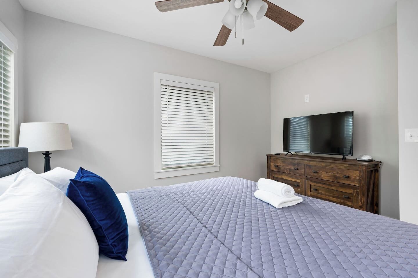 Ein Schlafzimmer mit Bett, Fernseher, Kommode, Deckenventilator und einem Fenster mit Jalousien.