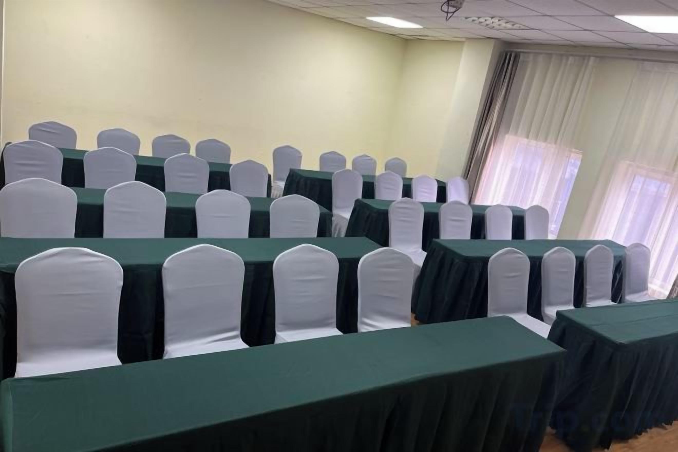 A conference room with rows of tables and chairs, a plain wall, and a window with curtains.