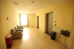 A hotel corridor with a seating area, a trash bin, a plant, and a door leading to another room. - Henghui Binle Hotel (Shijiazhuang Zhonghua North Street Branch) (Shijiazhuang)