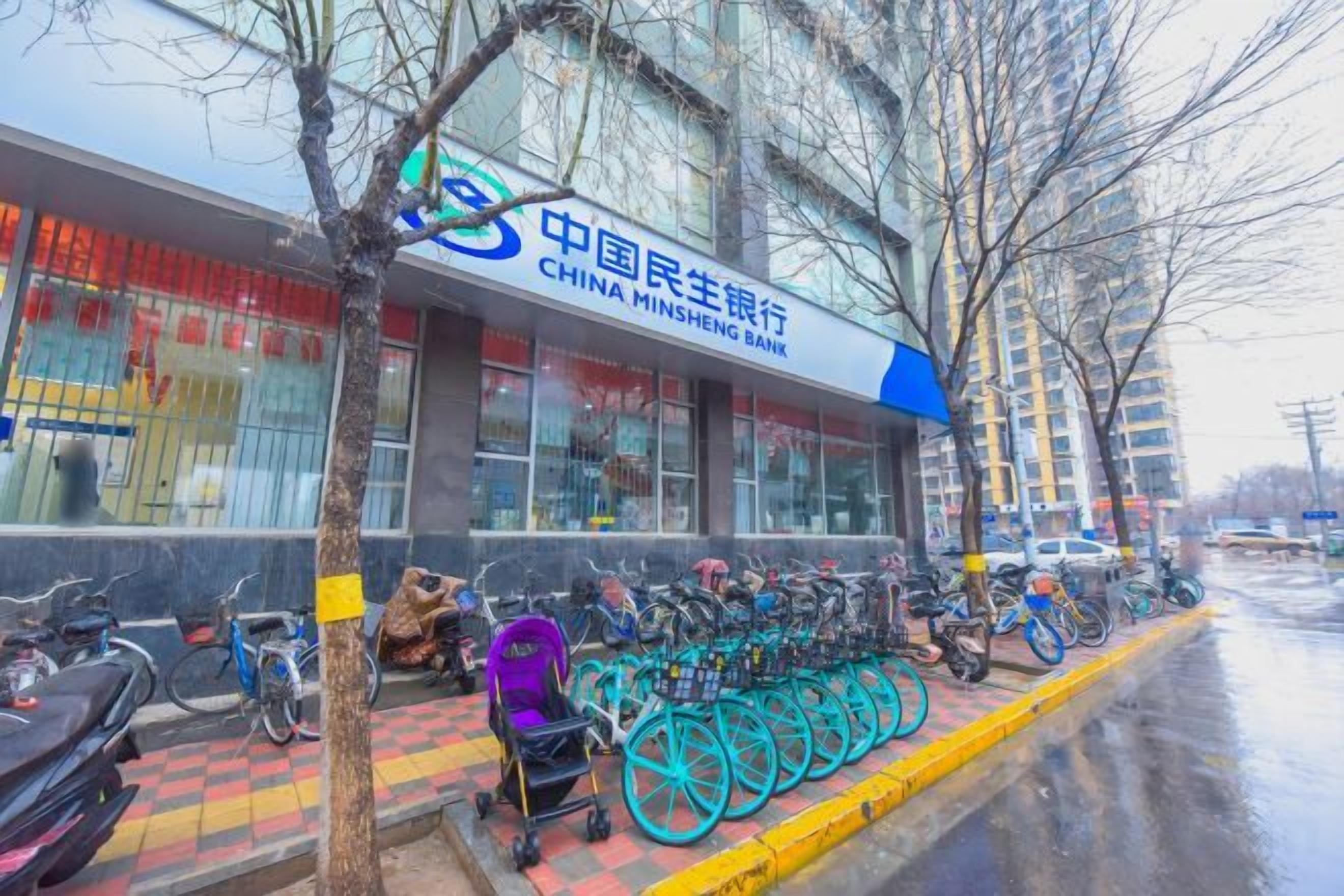 Una banca con insegna blu e bianca, biciclette parcheggiate fuori e un albero di fronte.