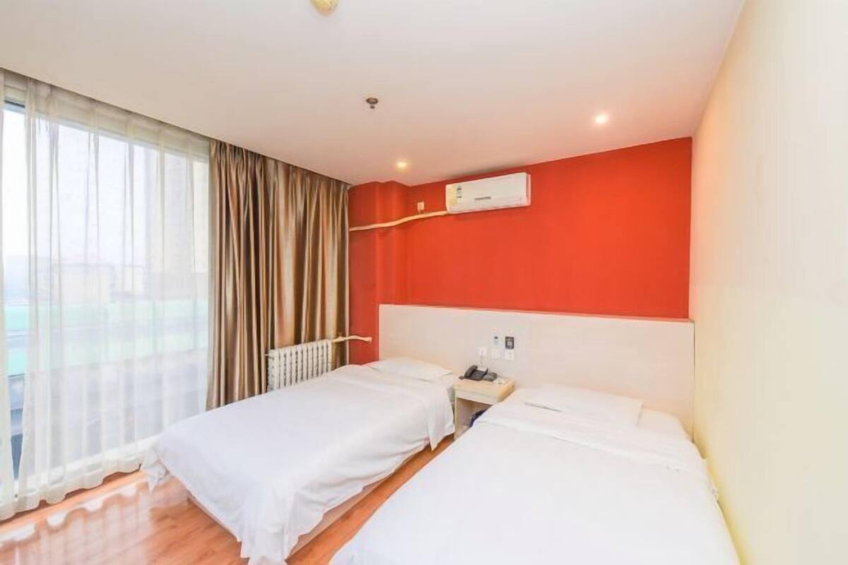 A hotel room with two beds, a window with curtains, an air conditioning unit, and a white bedside table.