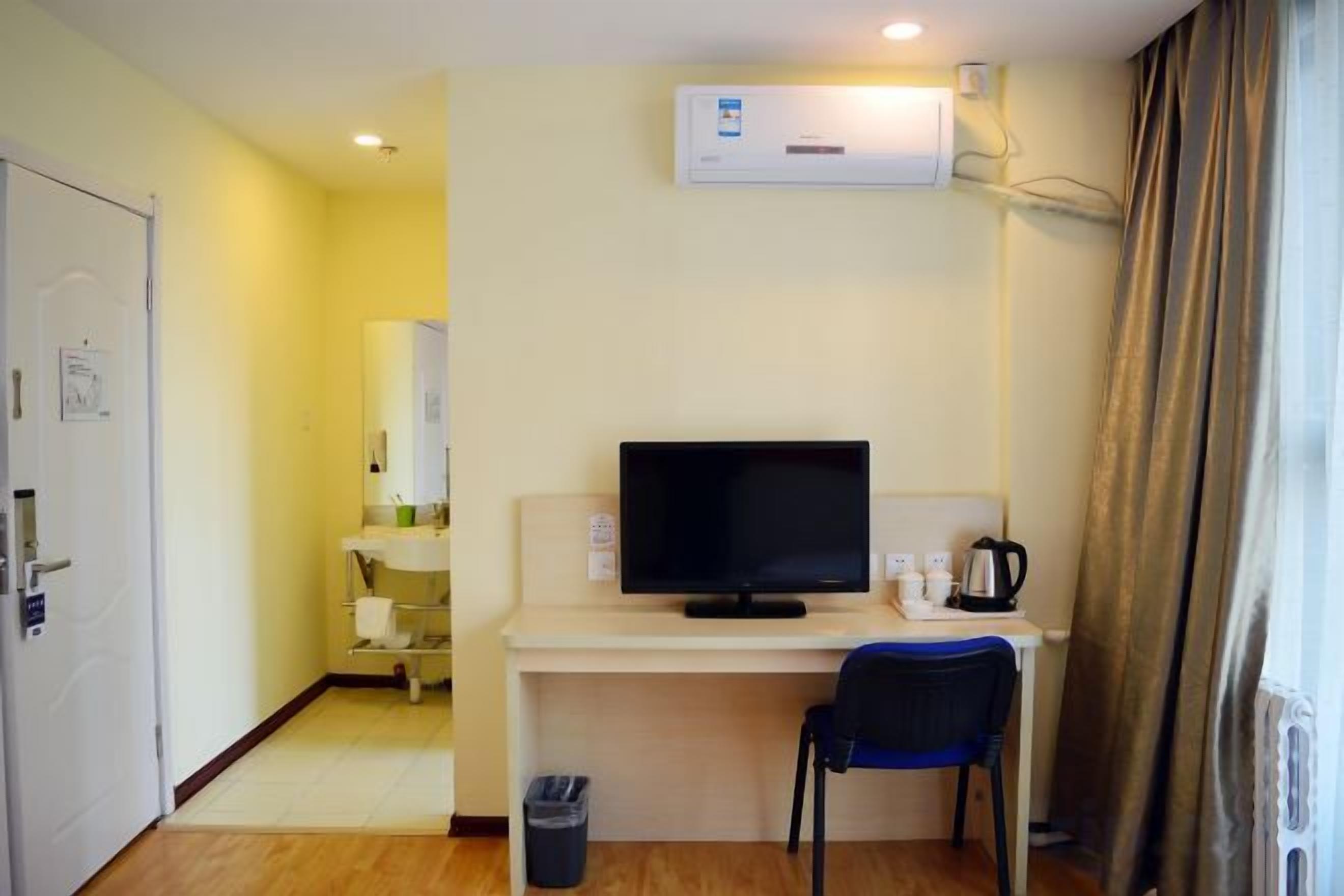 A hotel room with a television, a desk, a chair, a waste bin, and a window with curtains.